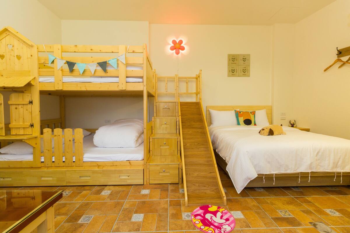 A room with a bunk bed, a single bed, a small wooden playhouse, and a wall-mounted light fixture.