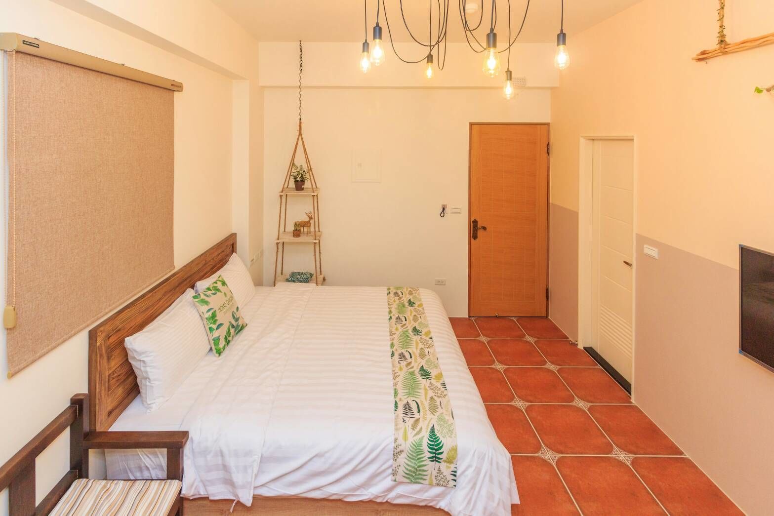 A bedroom with a bed, a wooden headboard, a hanging light fixture, a small wooden shelf with a plant, and a door.