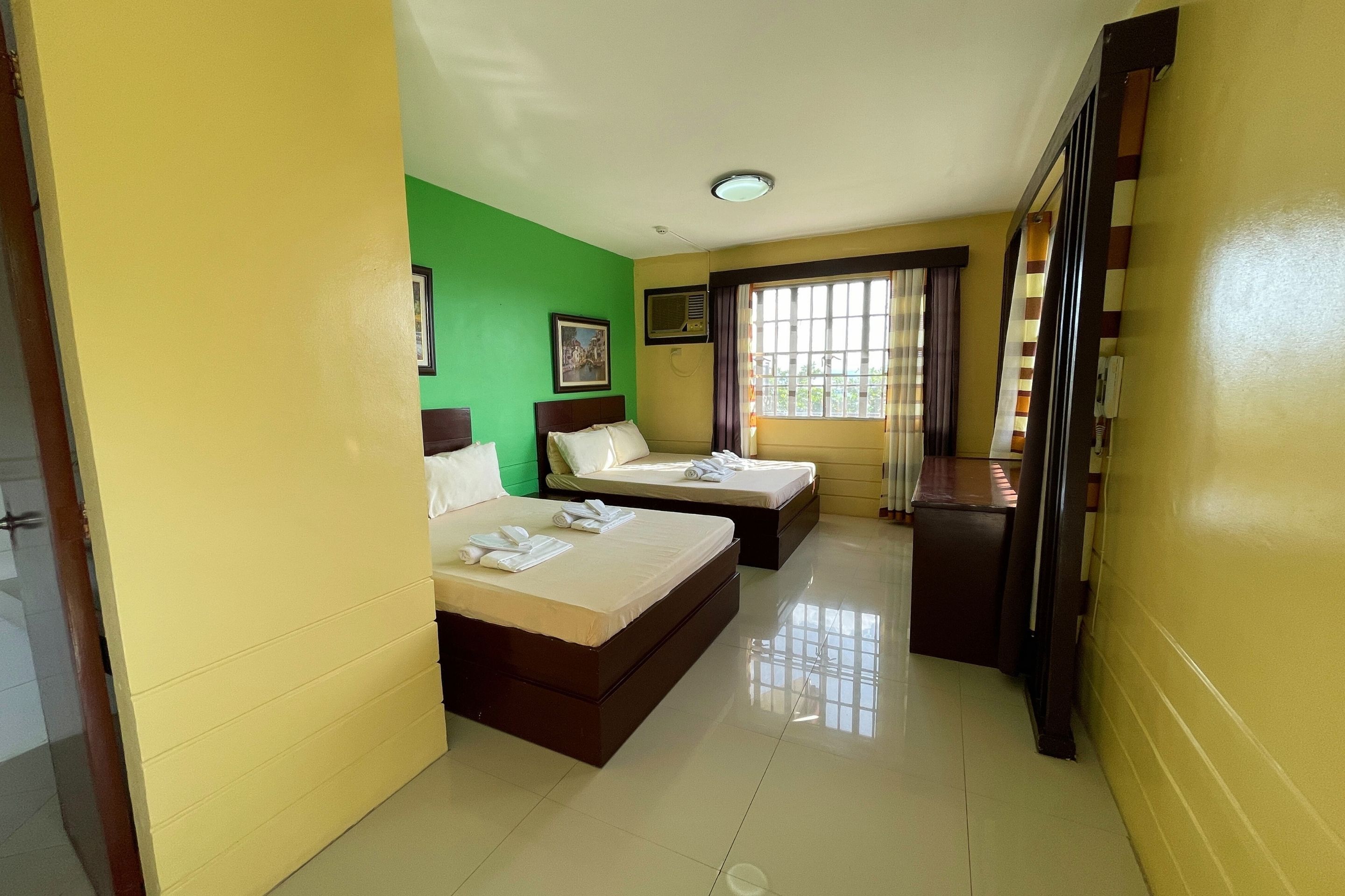 A hotel room with two beds, a small desk, a window with bars, and a green wall.