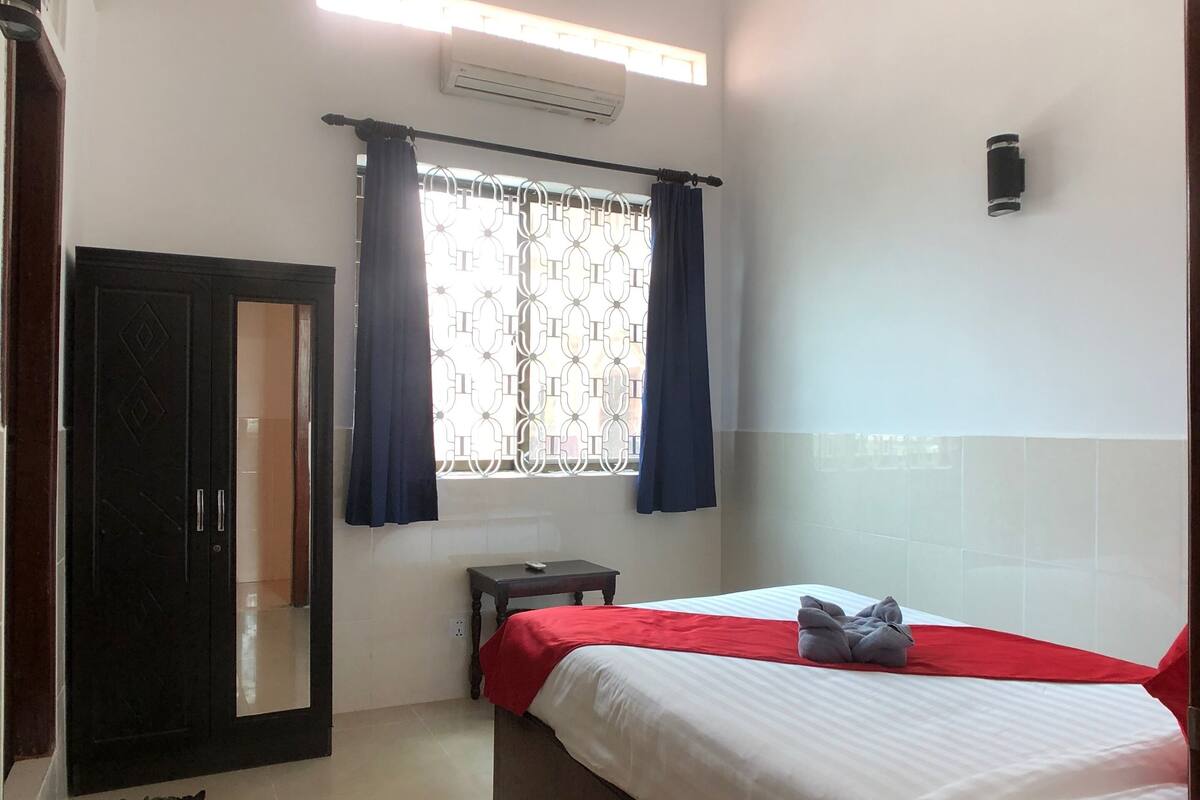 Een slaapkamer met een bed, een plafondventilator, een airconditioning, een raam met gordijnen, een nachtkastje en een kledingkast.