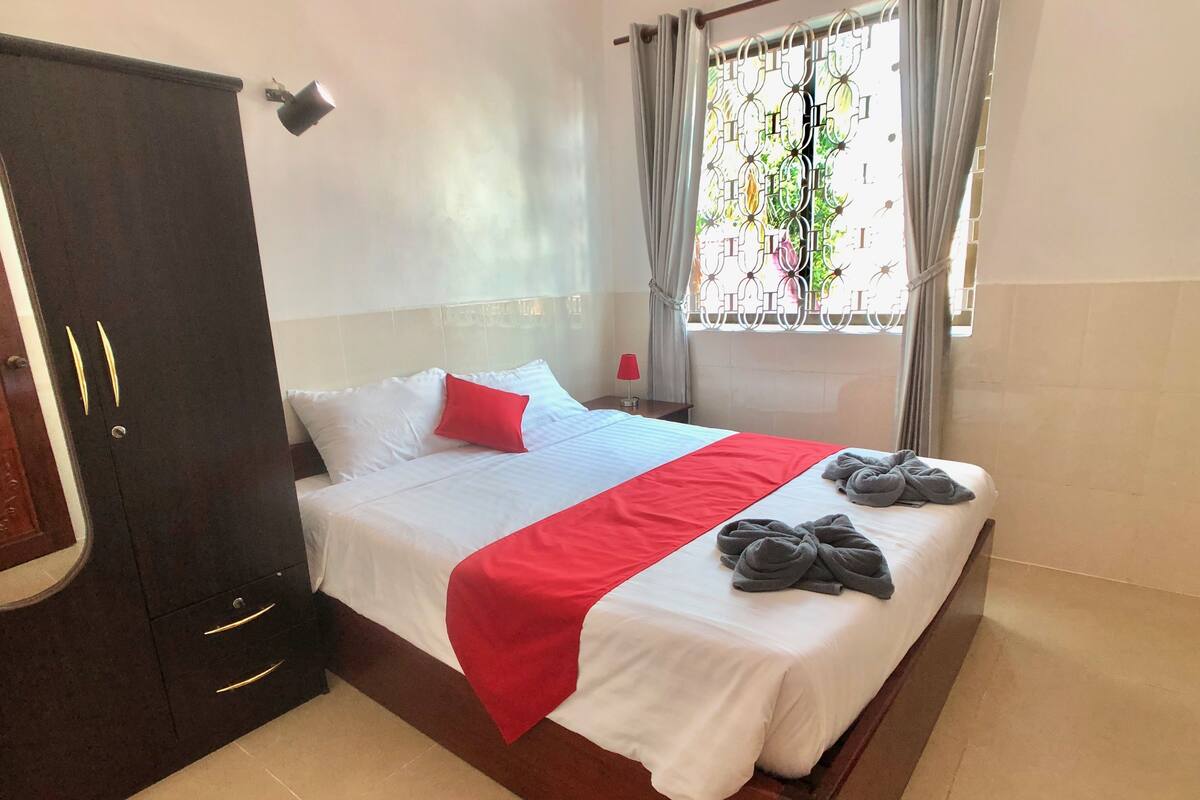 Een opgemaakt bed met wit en rood beddengoed, een rood kussen en twee handdoeken ernaast.