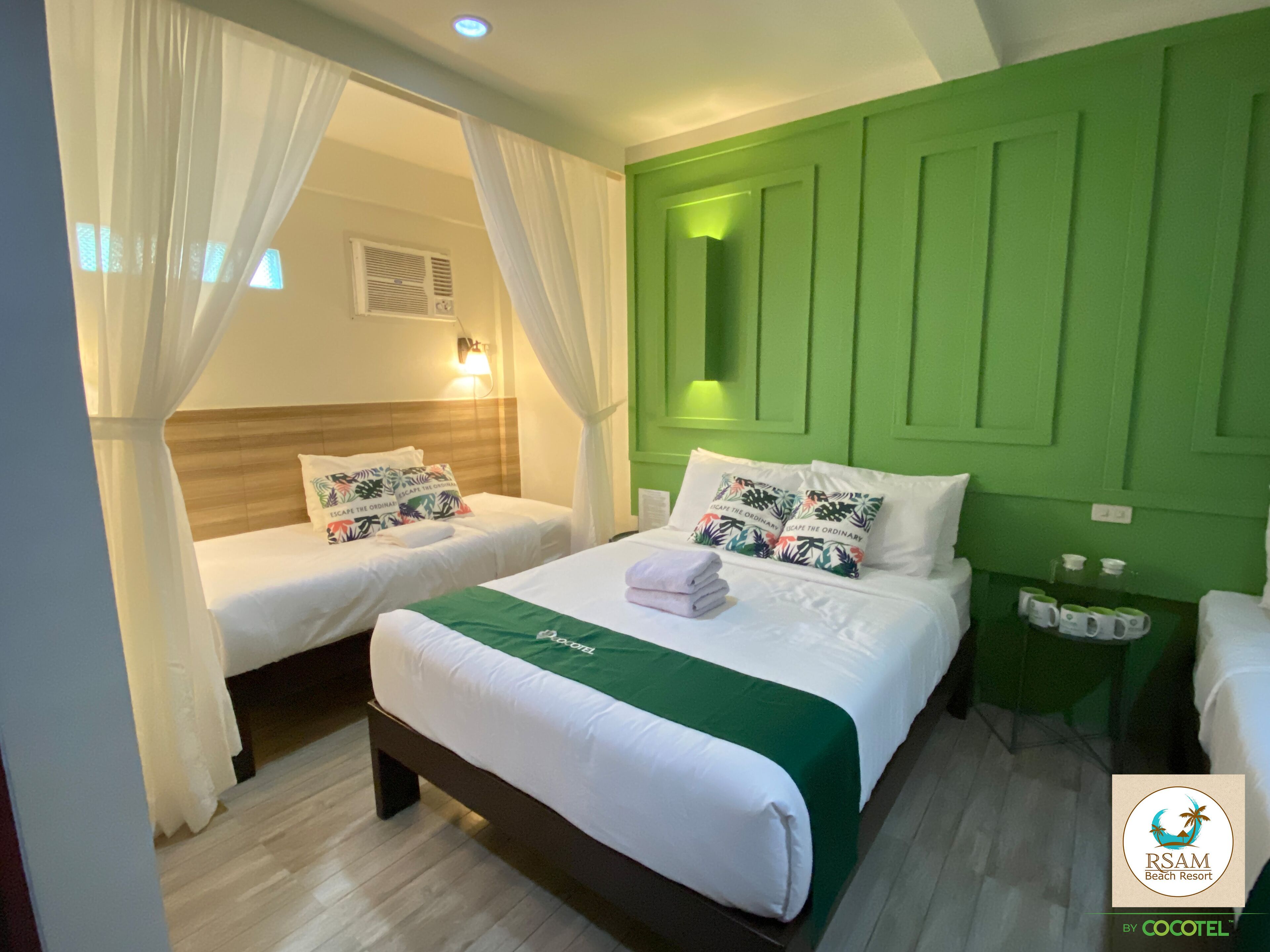 Habitación de hotel con dos camas, una pared de acento verde y una mesita de noche con lámpara.
