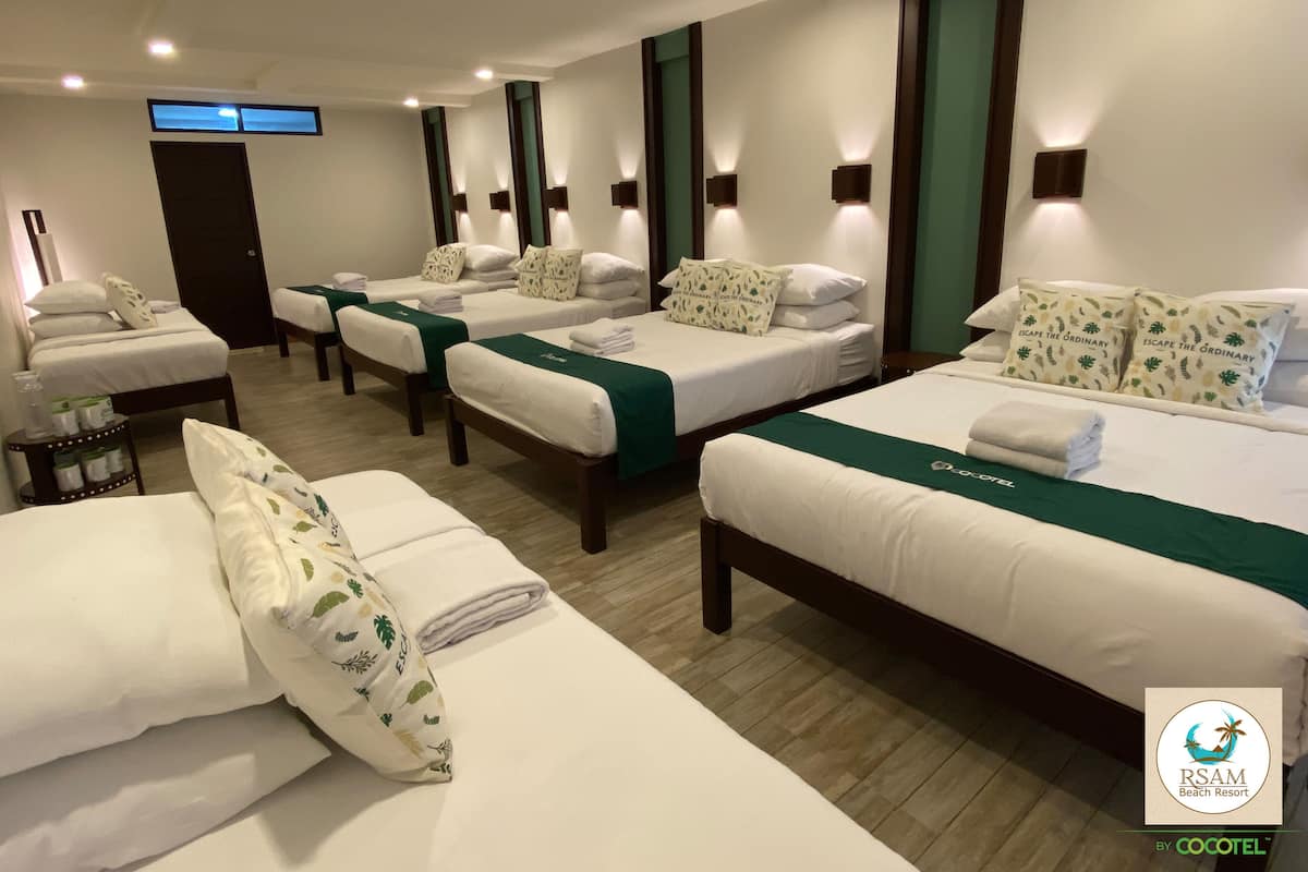 Ein Hotelzimmer mit vier Betten, jedes mit weißer Bettwäsche und grünen Kopfteilen. Es gibt Wandleuchten und einen Flachbildfernseher an der Wand.