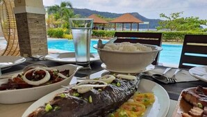 Uma mesa de jantar com diversos pratos, incluindo um peixe inteiro, arroz e outros alimentos, com uma piscina e montanhas ao fundo.