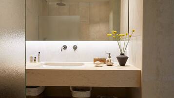 A modern bathroom with a large sink, a mirror, and a shower. There are baskets under the sink and a vase with yellow flowers on the countertop.