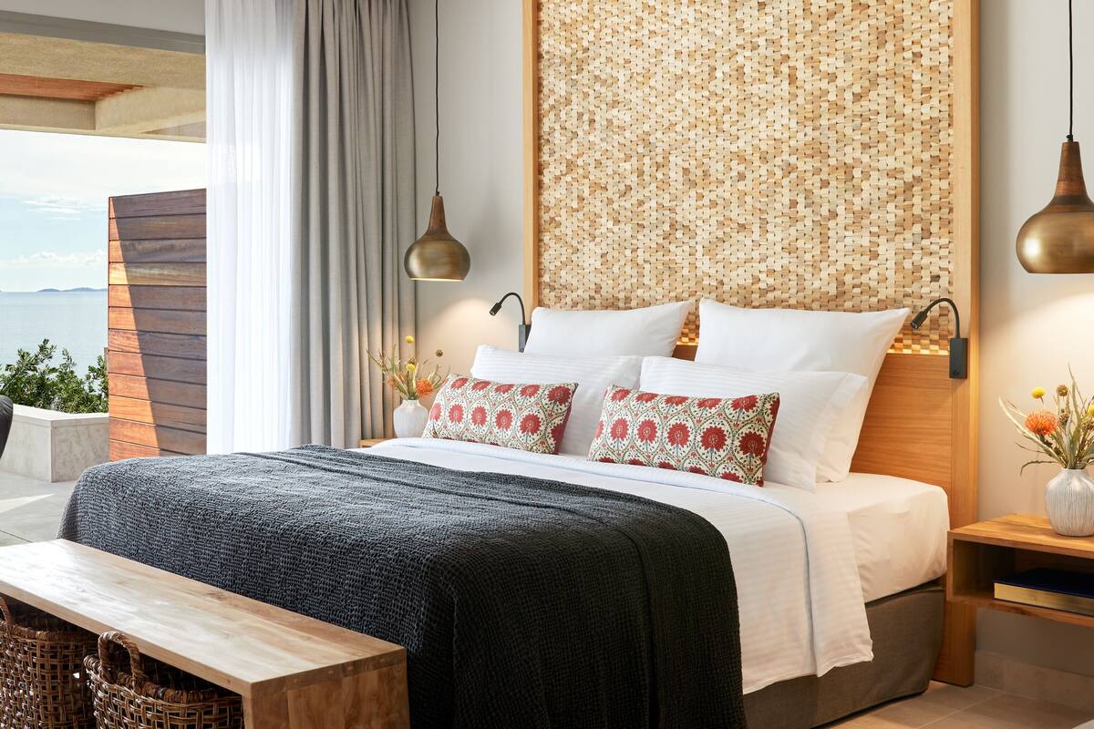 Un dormitorio moderno con una cama grande, cabecera de madera, vista al mar y una obra de arte tejida en la pared.