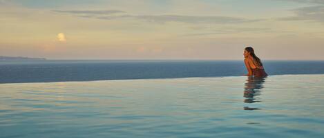 A woman standing in a infinity pool with a view of the ocean and a sunset.