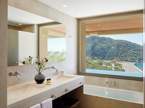 A modern bathroom with a large mirror, two sinks, a bathtub, and a view of a beach and mountains.