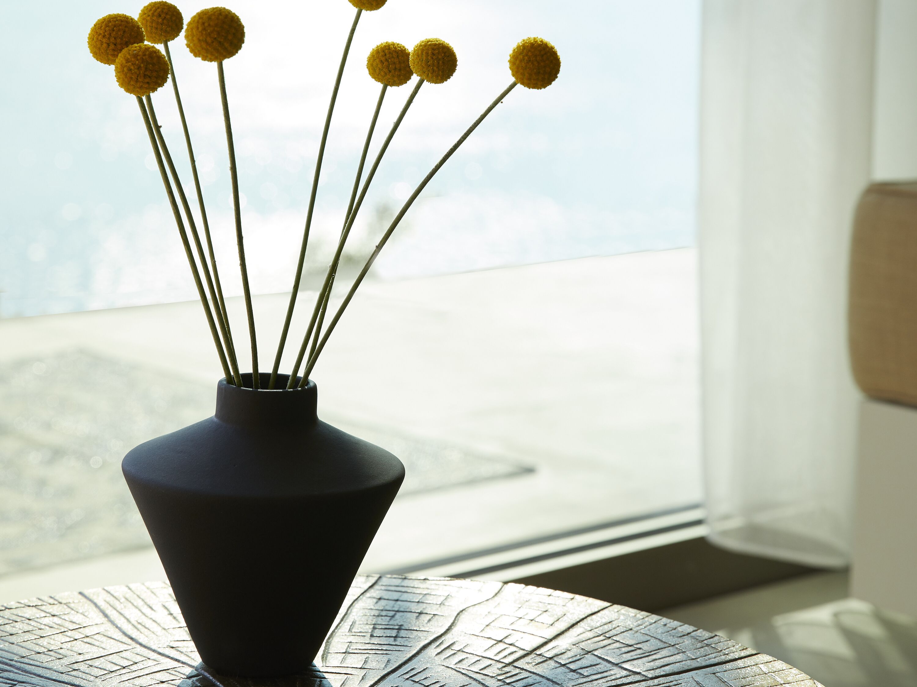 A black vase with yellow spherical flowers on a table by a window.