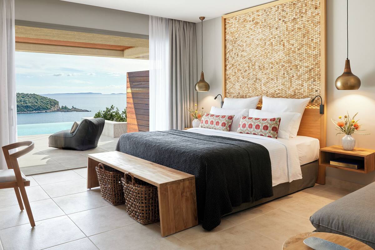 Un dormitorio moderno con una cama grande, vista al mar, cabecera de madera y un banco.