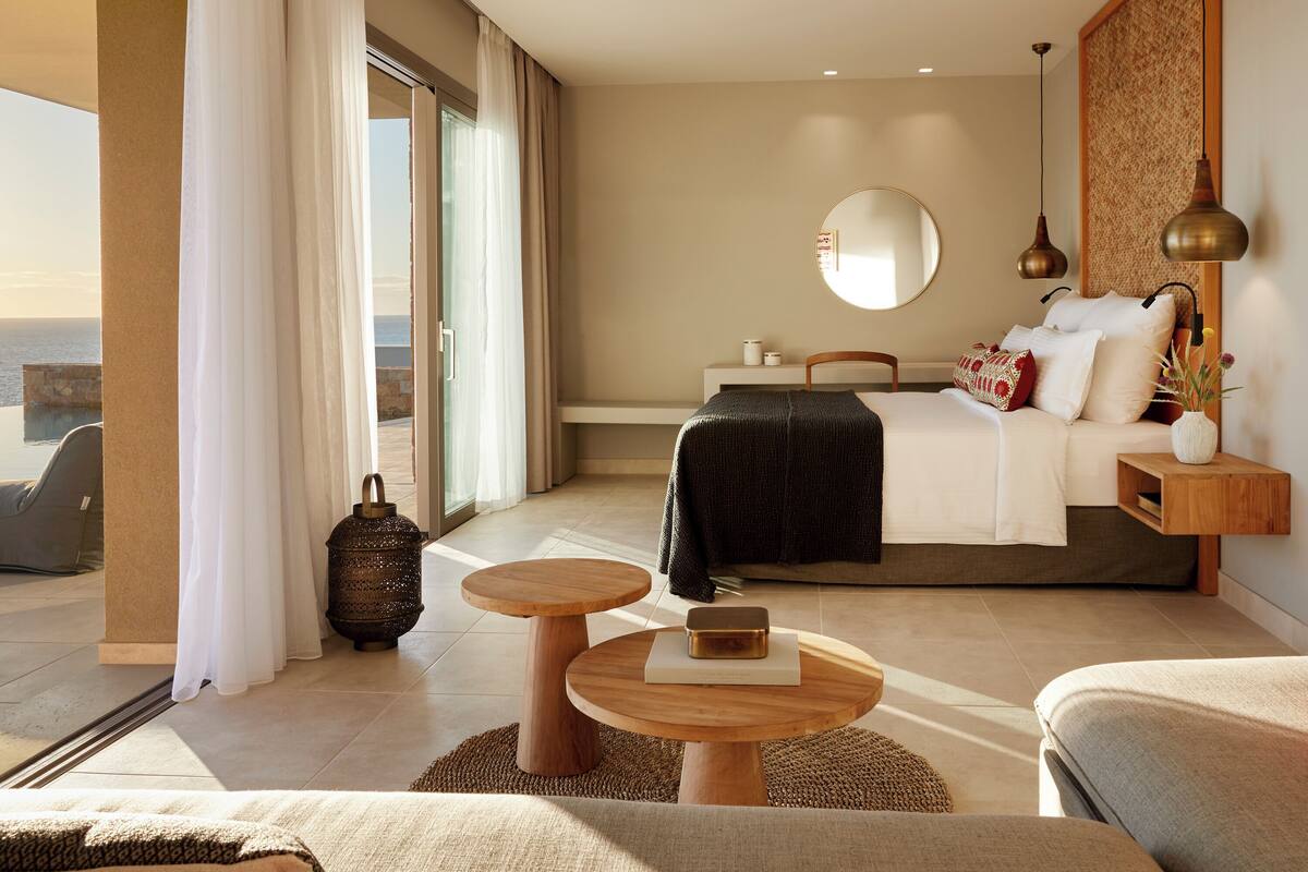 Una habitación de hotel moderna con cama, mesitas de noche, un espejo redondo y vista al mar.