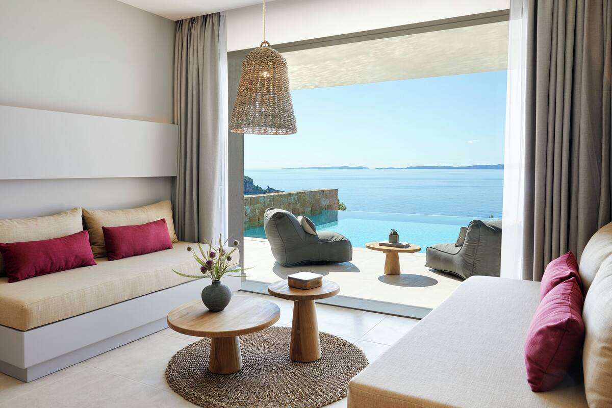 Una habitación de hotel moderna con sofá, sillones, una mesa de comedor y vista al mar.