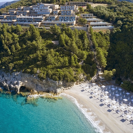 An aerial view of a coastal resort with multiple hotel buildings, a sandy beach with umbrellas, and a rocky shoreline.