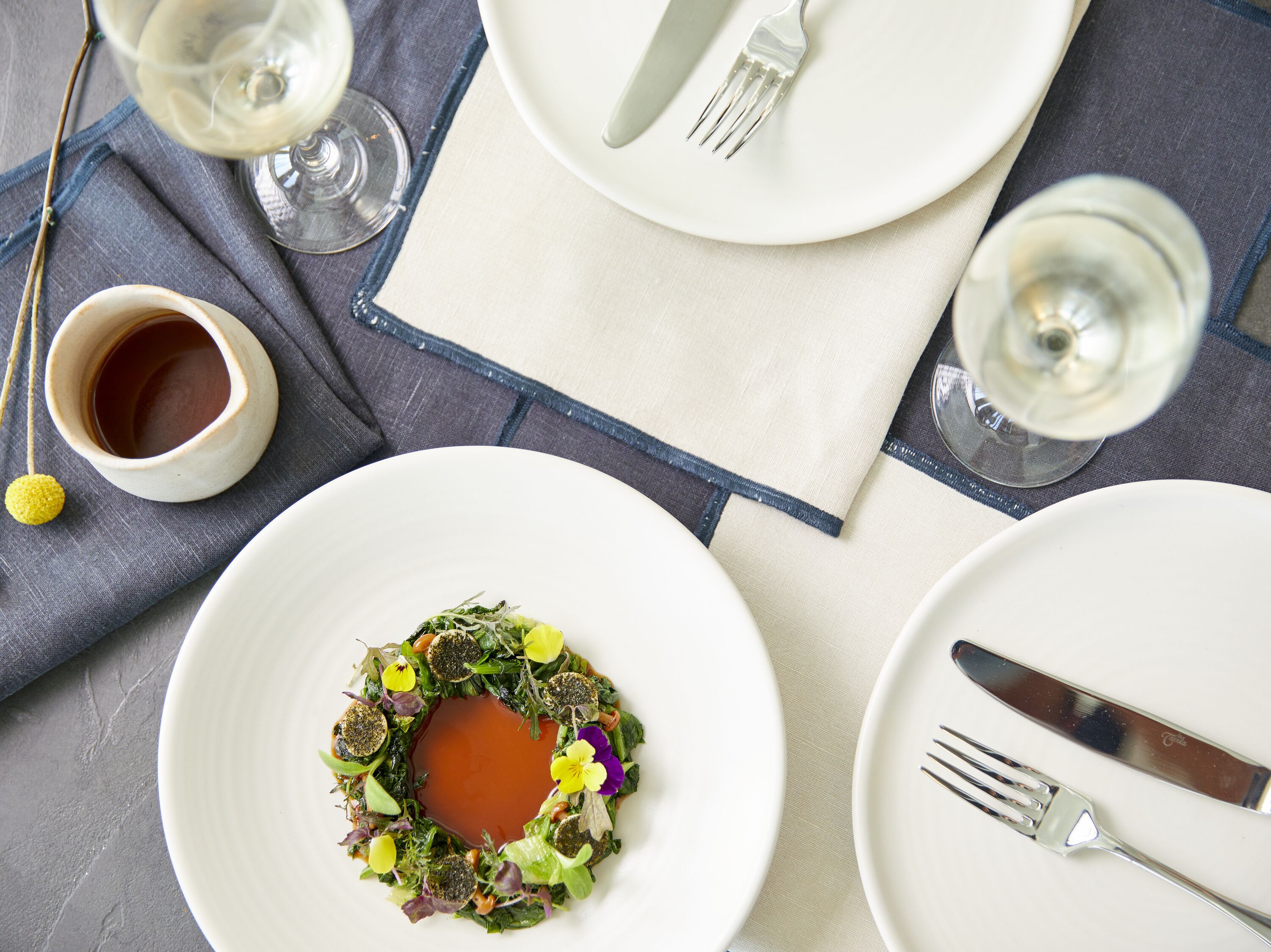 Une table dressée avec de l’argenterie, des assiettes blanches et un verre de vin blanc.