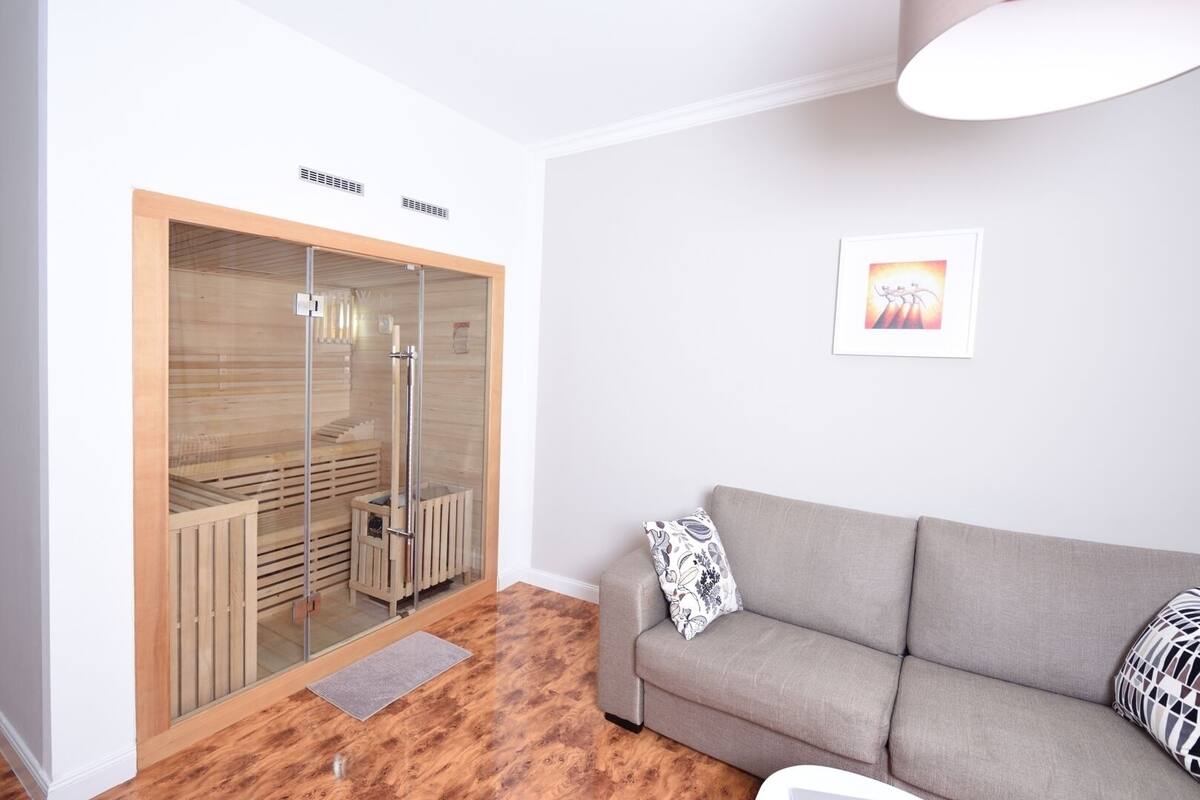 A modern living room with a grey sofa, a glass-walled sauna, and a framed picture on the wall.