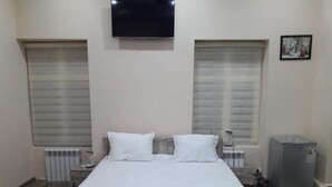 A bedroom with a bed, two windows with blinds, a television on the wall, and a small refrigerator. - Nozisayyoh (Bukhara)