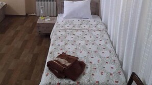 A small, neatly arranged bedroom with a single bed, a bedside table, a radiator, a window with blinds, and a picture on the wall. - Nozisayyoh (Bukhara)