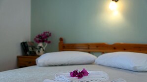 Una cama bien tendida con sábanas blancas y una flor morada, un cabecero de madera y una mesita de noche con un florero de flores.