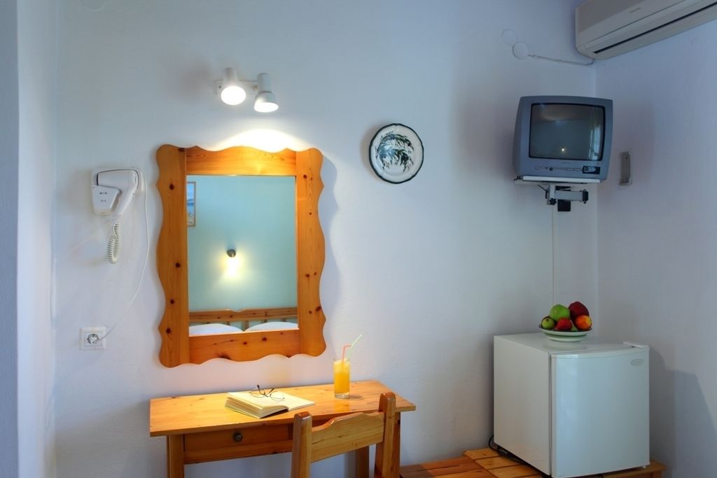 A small room with a wooden desk, a mirror, a small refrigerator, a telephone, and a television.