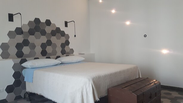 A bedroom with a bed, pillows, a bedside table, a wooden bench, a wall with hexagonal tiles, and wall-mounted lights. - Casa Maltese ri Scicli (Scicli)