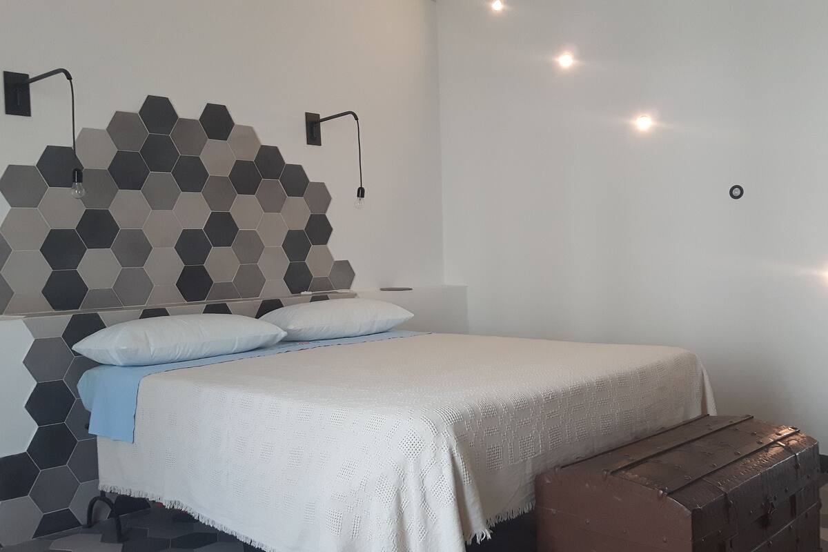 Quarto com cama, almofadas, mesa de cabeceira, banco de madeira, parede com azulejos hexagonais e luminárias de parede.