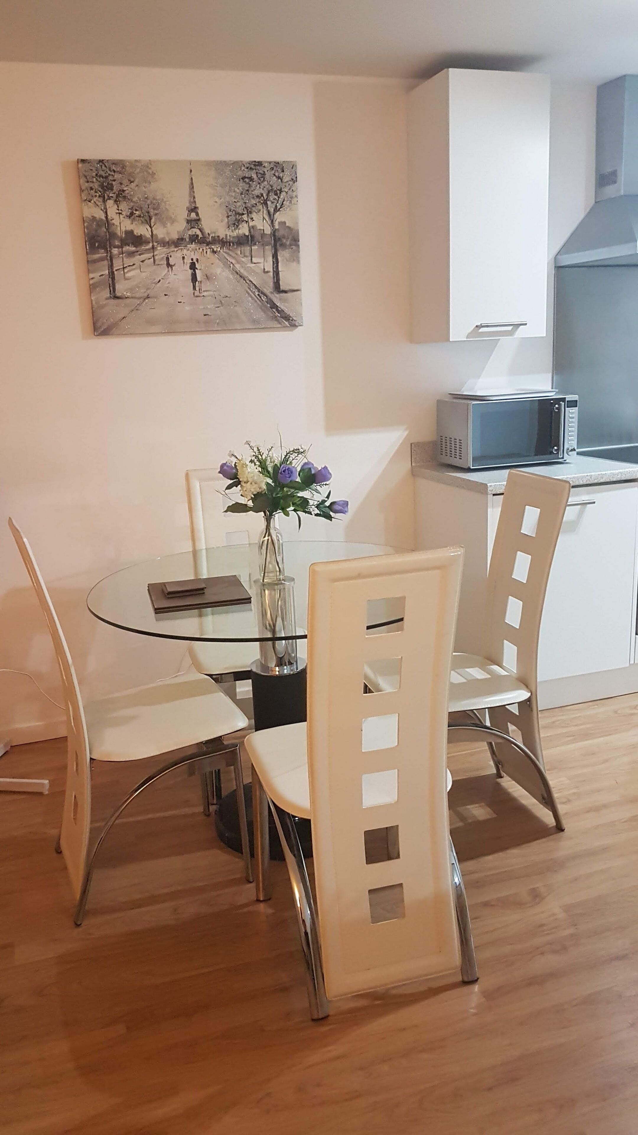 A dining area with a glass table, white chairs, a vase with flowers, and a painting of a street scene.