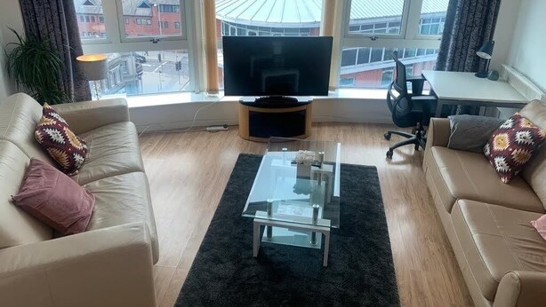 Moderni olohuone, jossa on lasinen sohvapöytä, beigen sävyiset sohvat, litteänäyttöinen televisio ja suuri ikkuna, josta avautuu näkymä kaupunkiin.