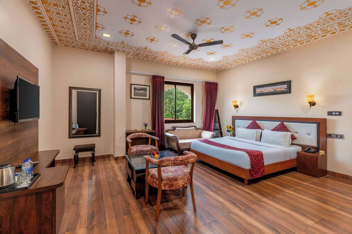 A hotel room with a bed, a desk, two chairs, a ceiling fan, and a TV.