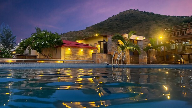 A swimming pool with a stone and wood structure in the background, surrounded by greenery and a mountain. - Vijaygarh Resort & Cottages with Private Pool (Udaipur)