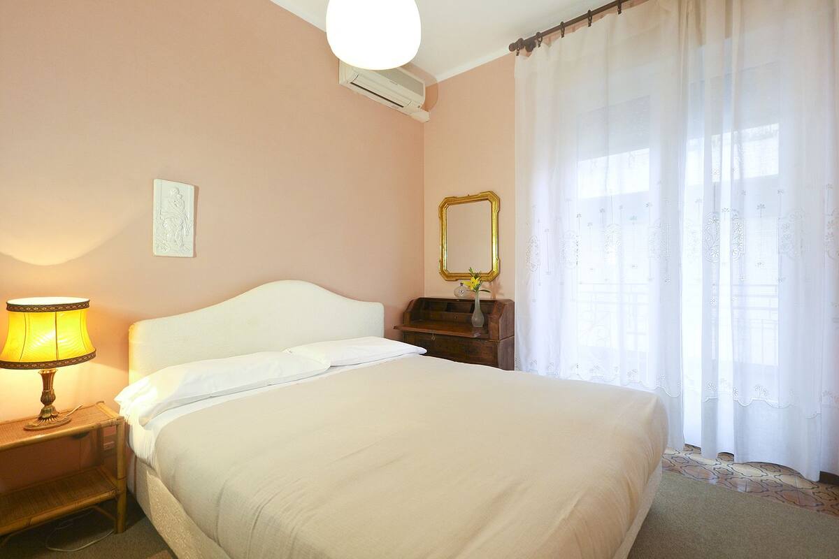 Un dormitorio con cama, mesita de noche con lámpara, cómoda, espejo y ventana con cortinas translúcidas.