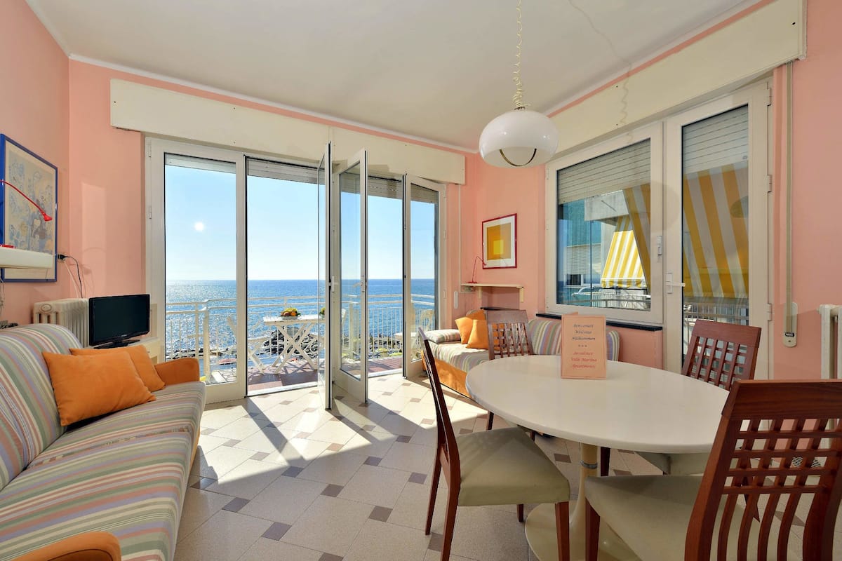 Una habitación con sofá, mesa de comedor y sillas, televisión y vista al mar a través de amplias ventanas.