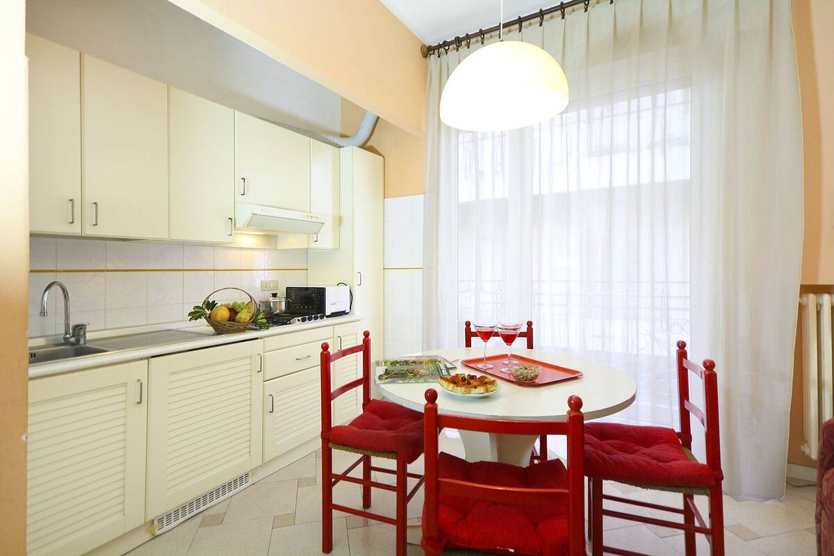Una cocina con armarios blancos, una mesa de comedor con sillas rojas y una ventana con cortinas de gasa.