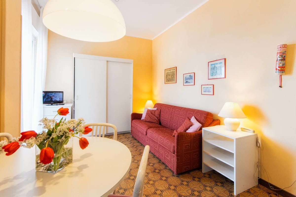 Una sala de estar acogedora con un sofá rojo, una mesita auxiliar blanca con una lámpara, una mesa de comedor redonda con un jarrón de flores y un televisor al fondo.