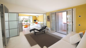 Een moderne woonkamer met een witte bank, een zwarte fauteuil en een bruin vloerkleed. Er is een glazen deur die leidt naar een balkon, waar een eethoek zichtbaar is.