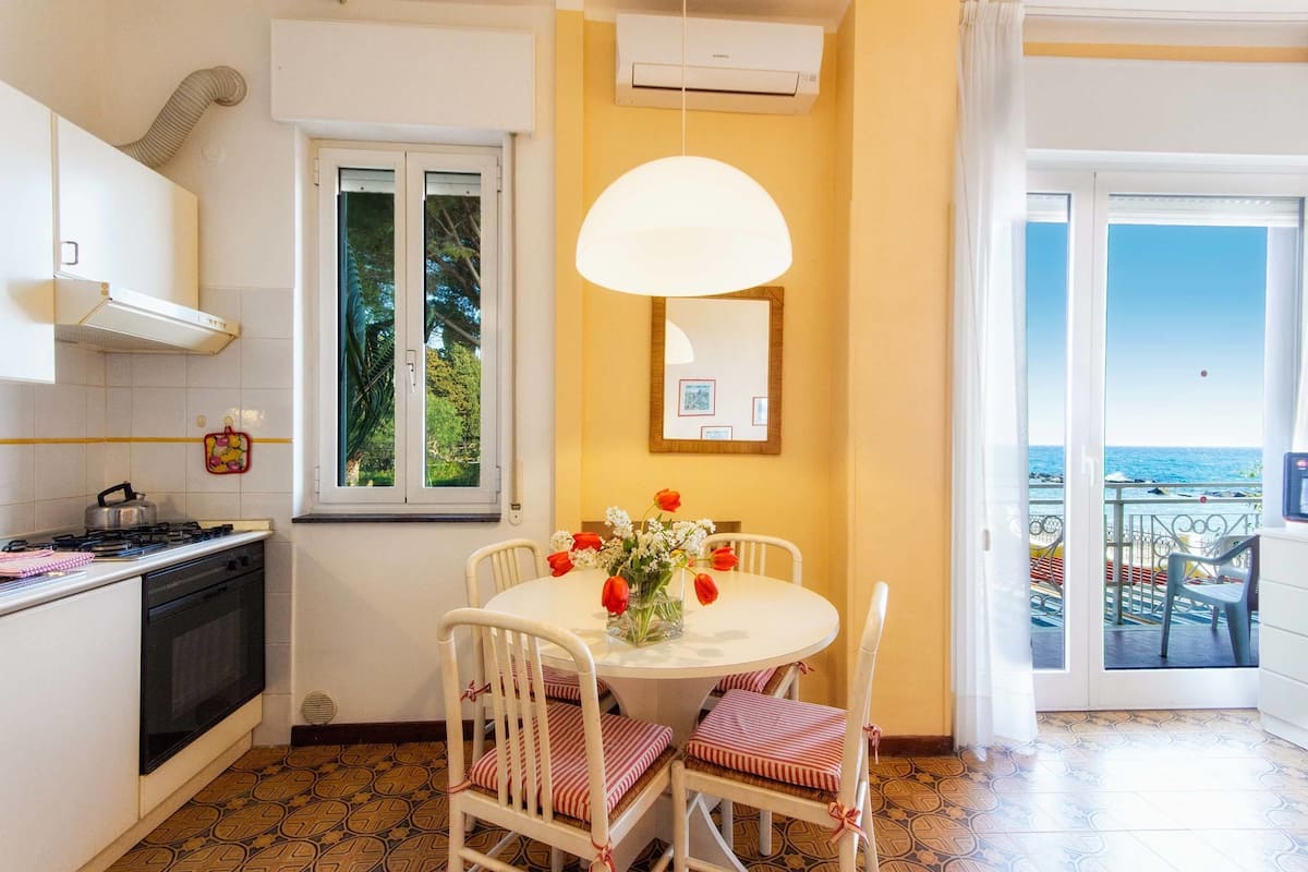 Una cocina con armarios blancos, una estufa y un comedor con mesa y sillas blancas. Se observa una ventana con vista al mar y un balcón con mobiliario exterior.
