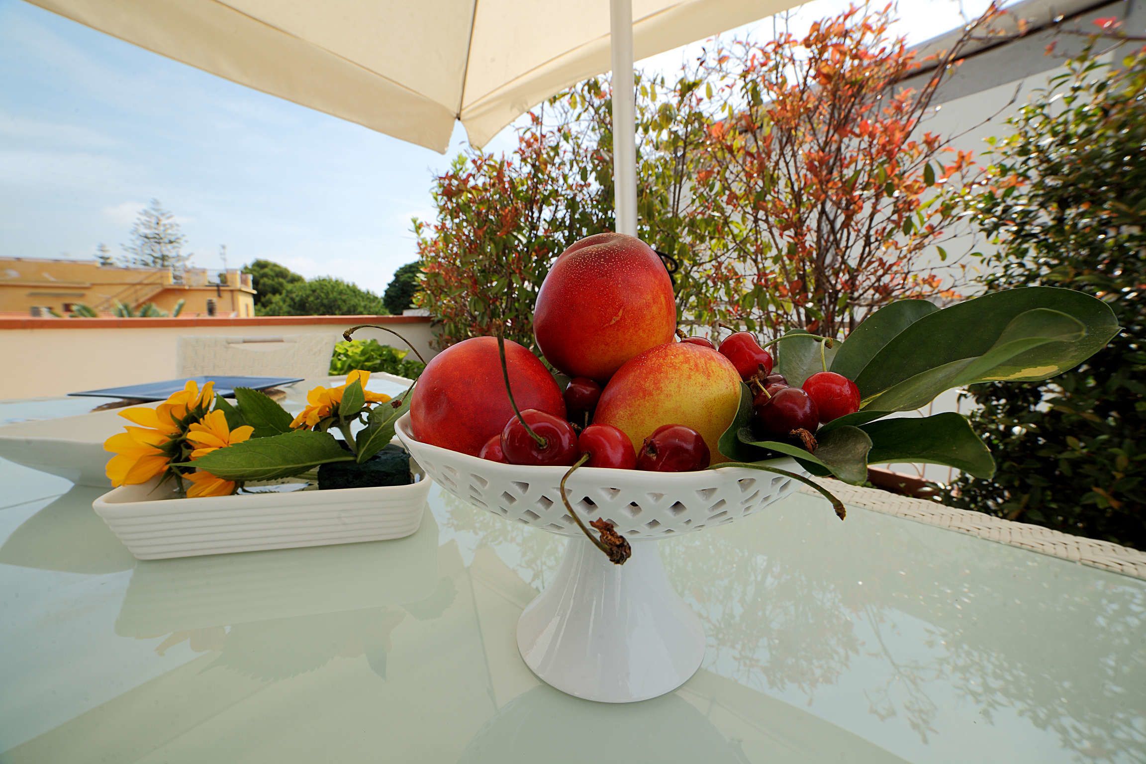 Un tavolo con una ciotola di frutta contenente mele e ciliegie, una piccola pianta in vaso con fiori gialli e un vaso bianco.