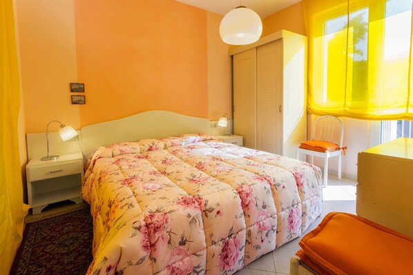A bedroom with a floral bedspread, a nightstand with a lamp, a chair, and a desk with a chair.