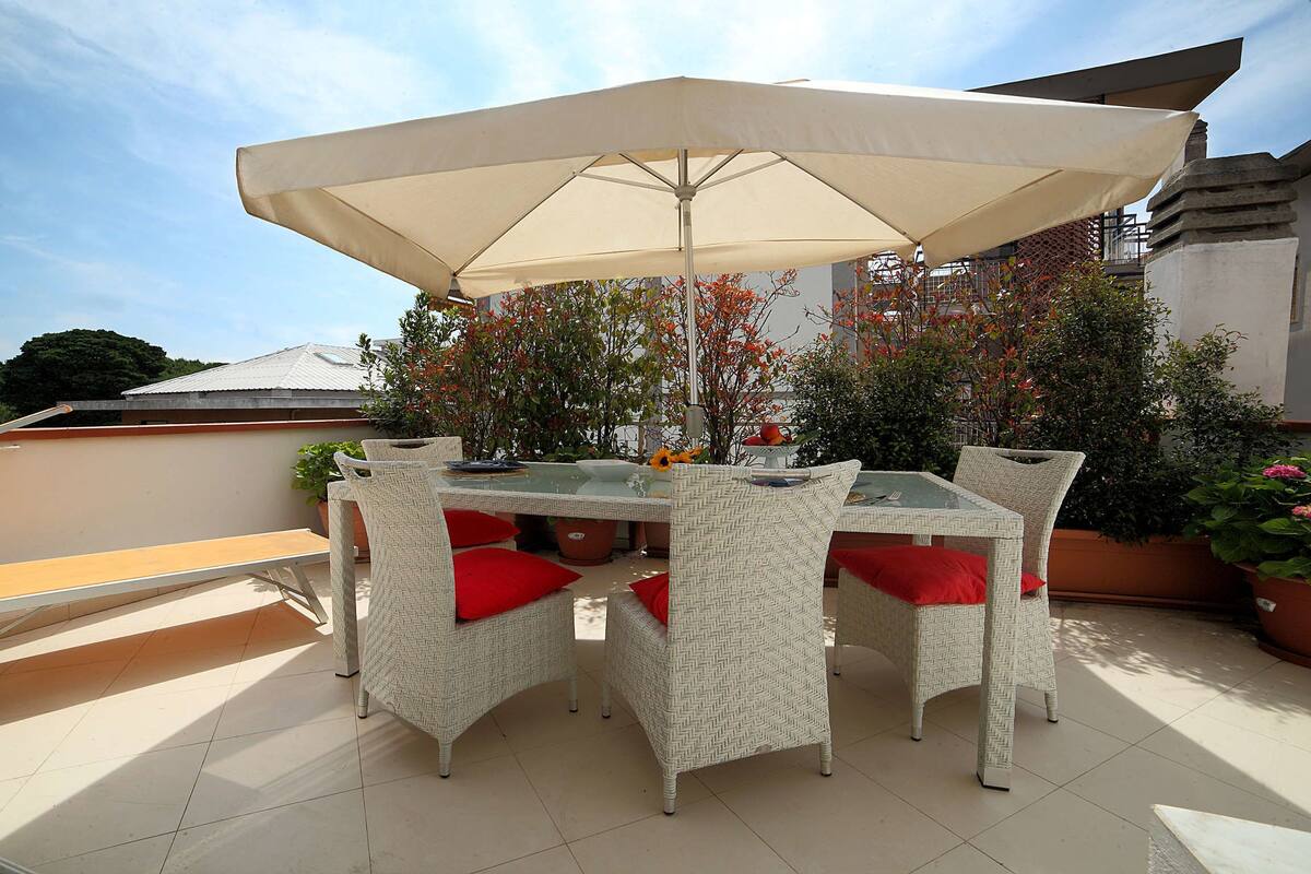 Un patio con una mesa de vidrio, sillas de mimbre, una sombrilla y plantas en macetas.