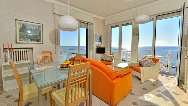 Een moderne woonkamer met een glazen tafel, een oranje bank en grote ramen met uitzicht op de zee.