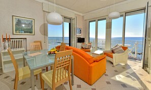 Een moderne woonkamer met een glazen tafel, een oranje bank en grote ramen met uitzicht op de zee.