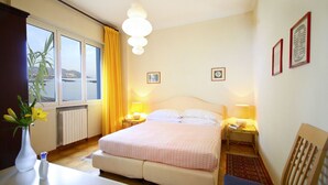 Hotelkamer met een bed, bedlampen, een raam met gordijnen, een radiator, een klein tafeltje met een bloemenvaas en een stoel.