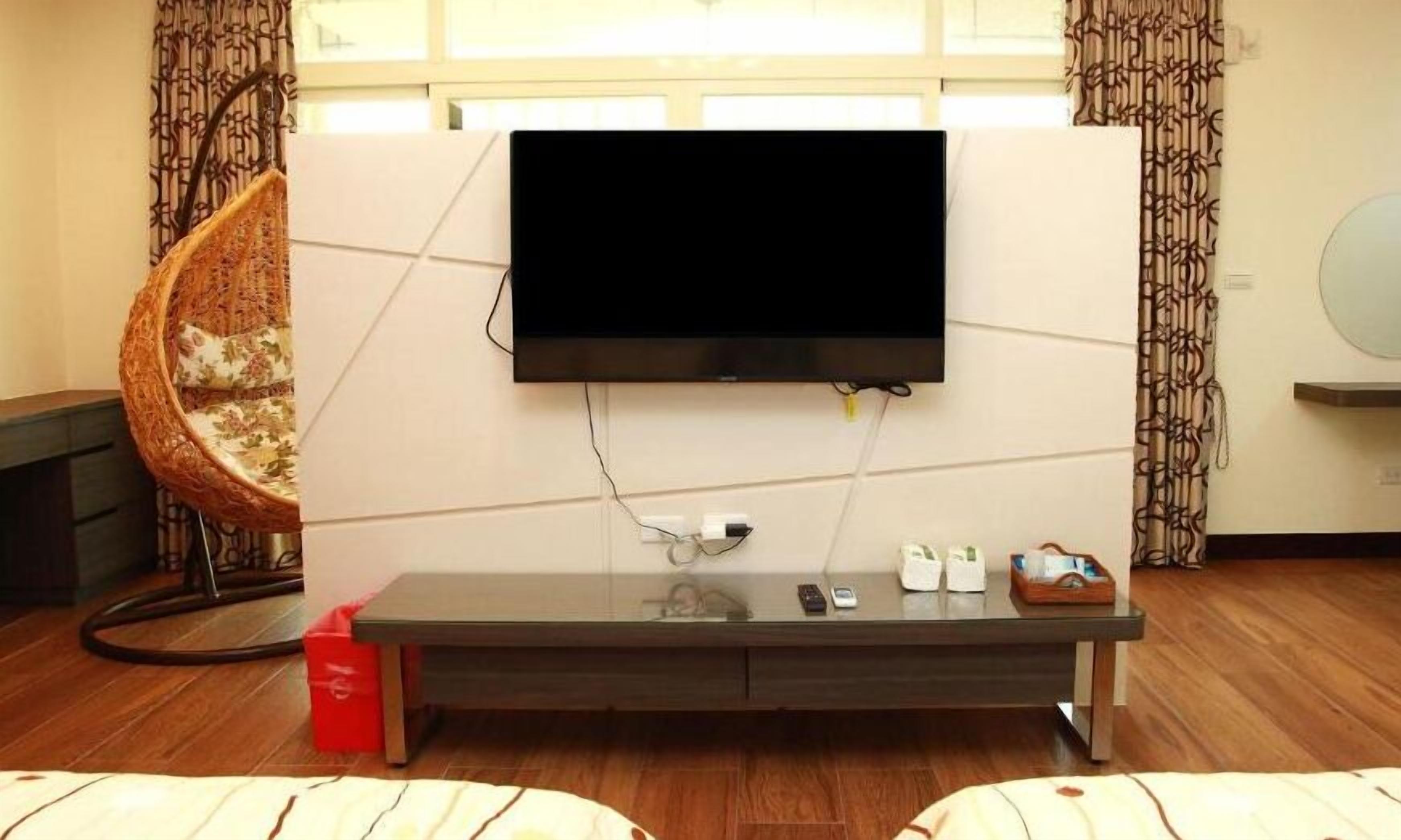 A modern living room with a flat-screen TV mounted on a wall, a glass coffee table, a wicker chair, and a wooden floor.
