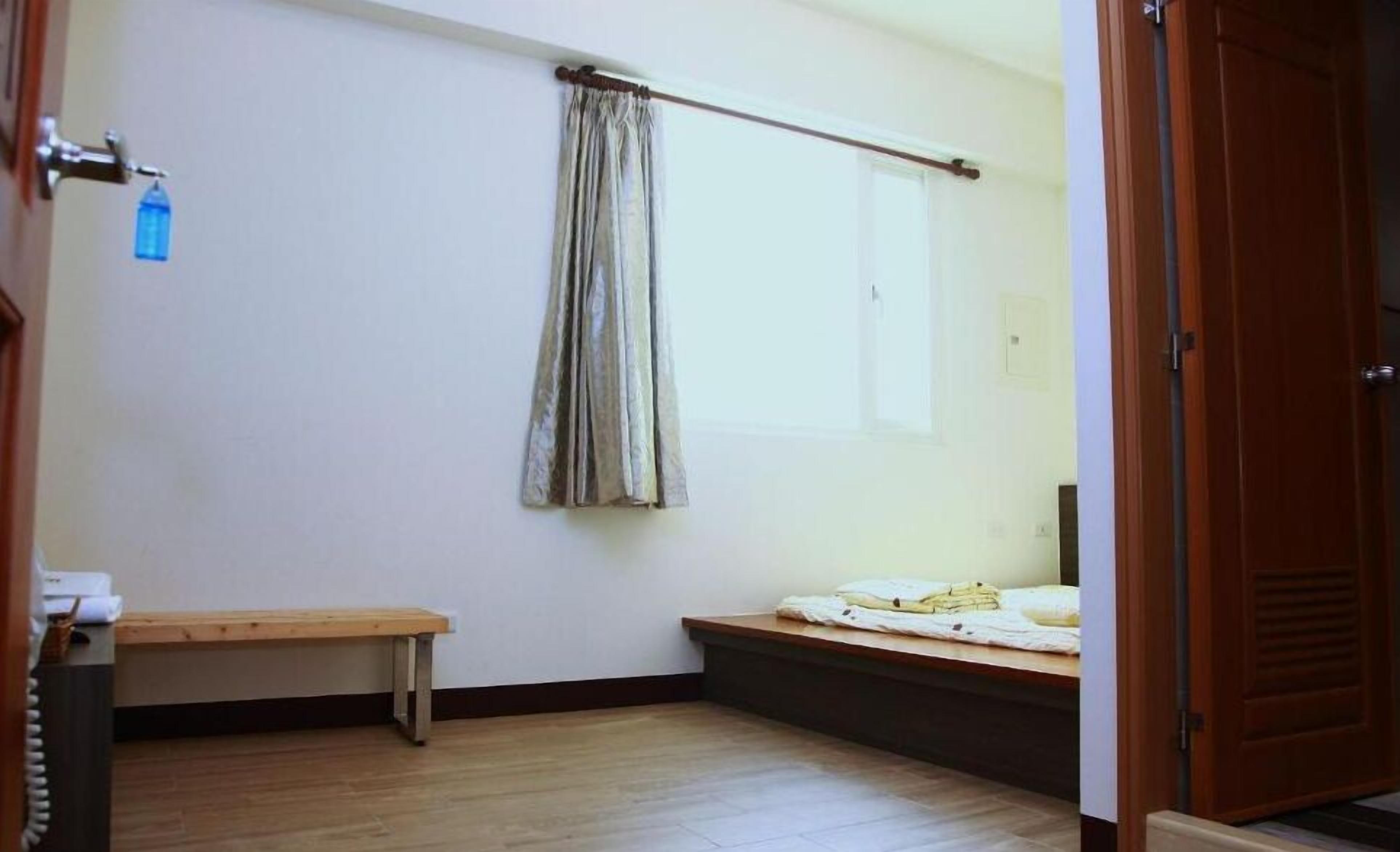 A small, sparsely furnished room with a wooden bench, a window with a curtain, and a door.