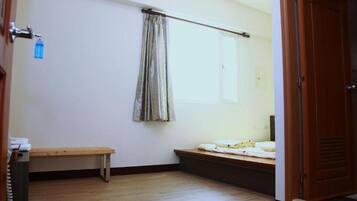 A small, sparsely furnished room with a wooden bench, a window with a curtain, and a door.