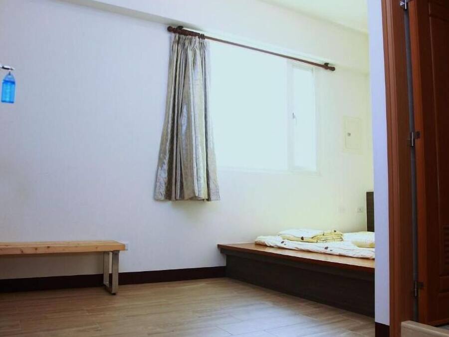 A small, sparsely furnished room with a wooden bench, a window with a curtain, and a door.