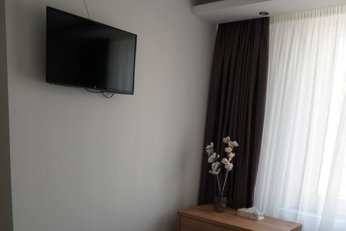 Habitación de hotel con cama, mesita de noche de madera, televisor de pantalla plana en la pared y ventana con cortinas.