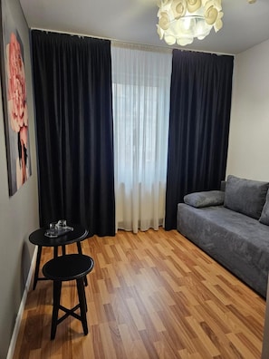 A room with a grey sofa, a small round table with two stools, a window with sheer and dark curtains, and a ceiling light fixture. - Meteor Central (Cluj-Napoca)