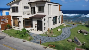 Un edificio moderno de dos pisos con un sendero de piedra que lleva a la entrada, ubicado cerca de una playa con el océano al fondo.