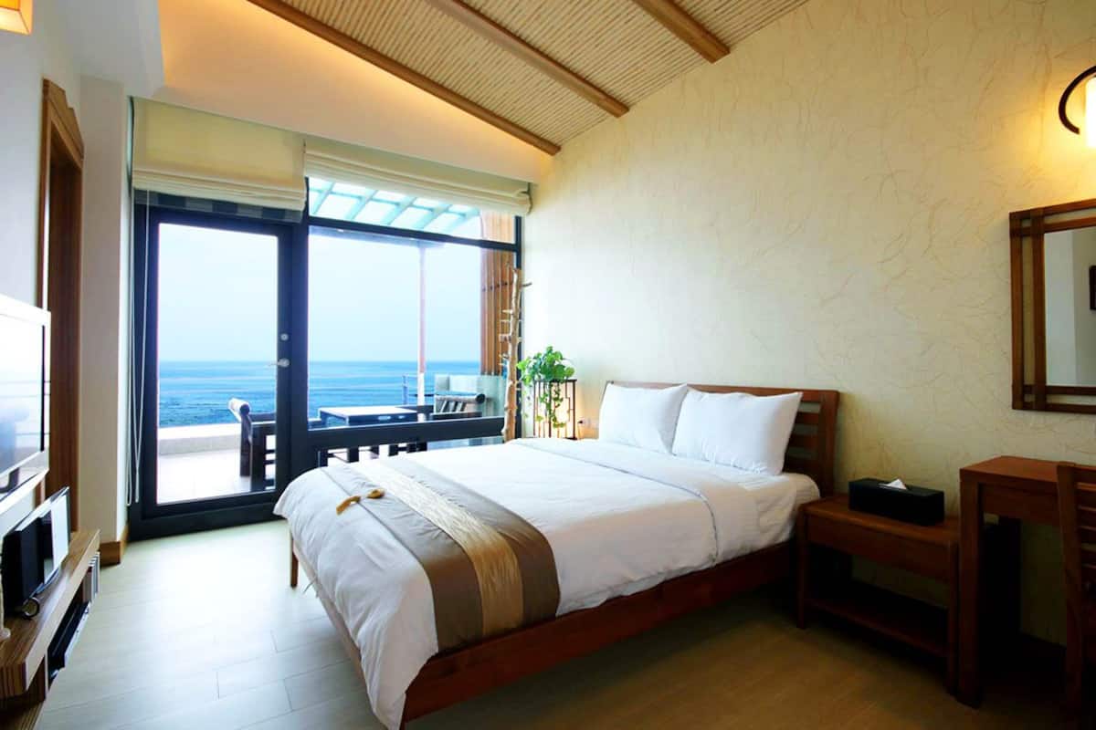 A hotel room with a large bed, a TV, a desk, and a balcony with a view of the ocean.