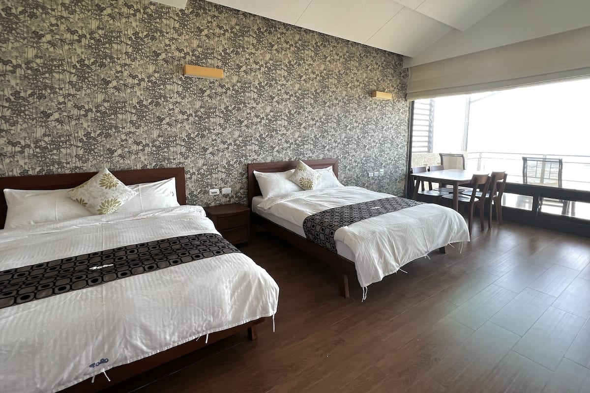 Two beds with wooden frames and white bedding, a large window with a view, and a wooden table with chairs.